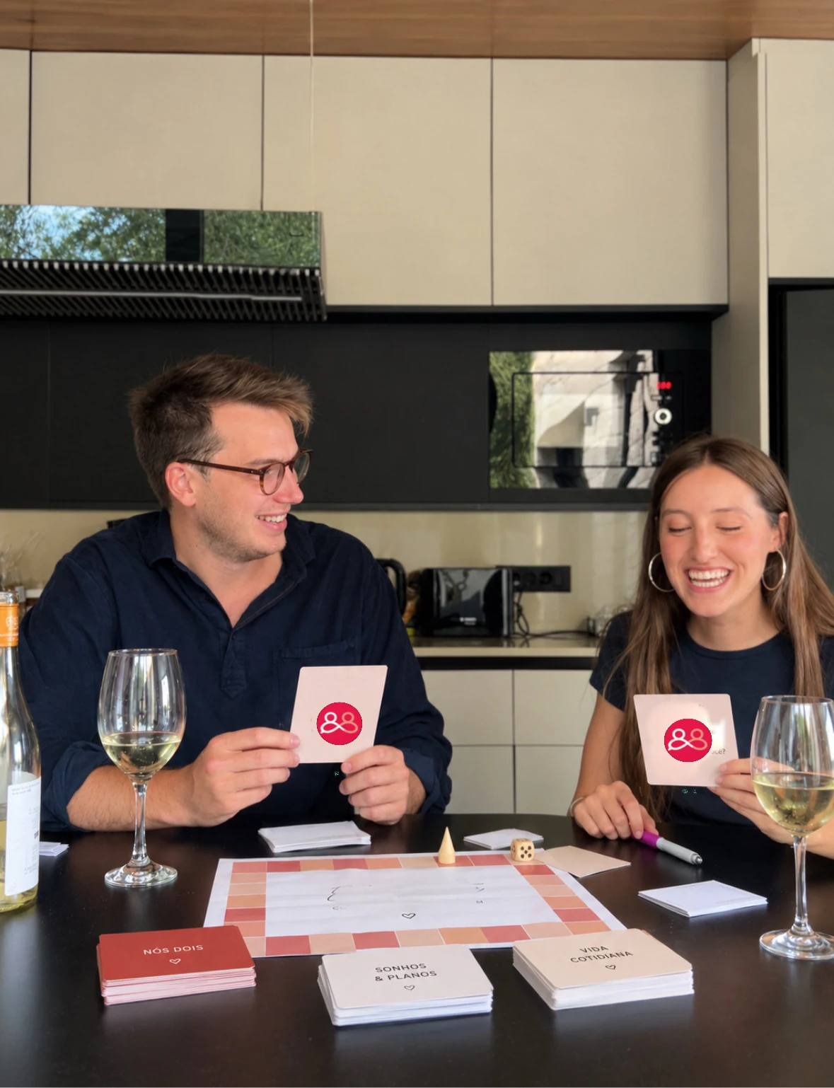 Casal rindo à mesa de casa vivendo a experiência Entre Gente que se Ama com cartas, dado e taças
